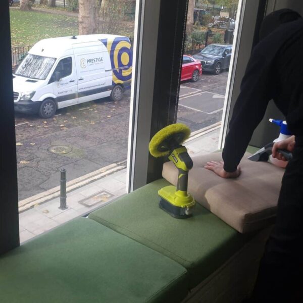 A Prestige technician cleaning first-floor upholstery at Canva, with the branded Prestige van visible through the window in Hoxton Square.