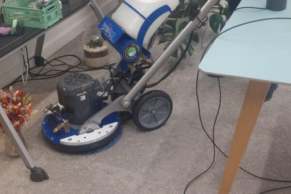 Prestige technician using an Orbot low-moisture machine to clean office carpet tiles near electrical floor boxes at Canva, Hoxton Square, London.